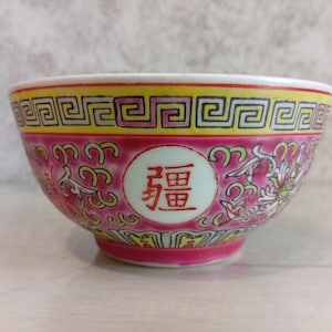 以下が含まれることがあります： ピンク色の外観の装飾的なセラミックボウル。花柄とスクロール模様が特徴です。縁は幾何学模様の黄色です。中央のメダリオンには赤い漢字が表示されています。