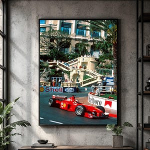 May include: A framed wall art print depicting a bright red Formula One race car driving on a track in Monaco. The scene features a grand building with arched architecture in the background, spectators on a staircase, and Shell and Marlboro signage.