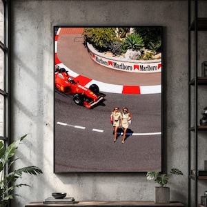 May include: A framed wall art print depicting a red Formula One race car navigating a curved track with a white and red curb. The track barrier features the text Marlboro. Two individuals in light-colored outfits walk across the asphalt.