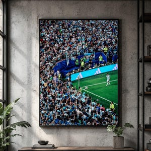 May include: A framed vertical wall art print depicting a high-angle view of a crowded soccer stadium. A lone player in a white jersey stands on the bright green grass near a corner flag, while a massive crowd in light blue and white fills the stands.
