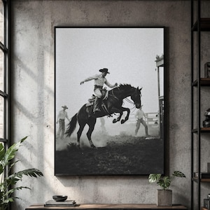 Könnte beinhalten: Ein schwarz-weiß gerahmter Kunstdruck, der einen Cowboy auf einem bockenden Pferd in einer staubigen Arena zeigt. Zwei weitere Personen mit Cowboyhüten stehen im Hintergrund in der Nähe einer Holzkonstruktion und fangen eine Rodeo-Szene ein.