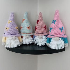 May include: Four crocheted gnomes with butterfly designs on their hats. The gnomes are in different colors: purple, light blue, pink, and pink. They are sitting on a black shelf.