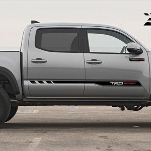 Op de afbeelding: Een zilveren pick-up truck met zwarte en rode "TRD Sport" stickers aan de zijkanten. De truck heeft zwarte wielen en een zwarte laadbak.