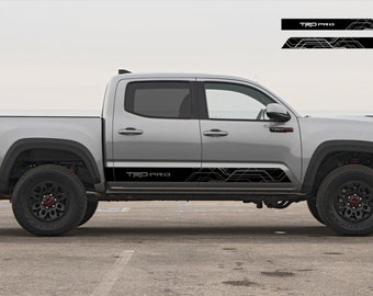 Könnte beinhalten: Ein silberner Pick-up mit schwarzen und weißen Grafiken an der Seite. Die Grafiken zeigen "TRD PRO" und ein geometrisches Muster. Der LKW hat schwarze Räder und Reifen.