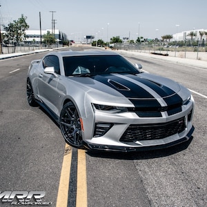 May include: A silver Chevrolet Camaro with black racing stripes on the hood. The car is parked on a paved road with yellow lines.