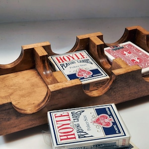 May include: A wooden card holder with three slots for holding decks of playing cards. The holder is made of light brown wood and has a natural finish. Two decks of Hoyle playing cards are visible in the holder, one deck is red and the other is blue.