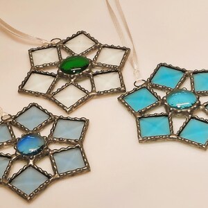 May include: Three stained glass snowflake ornaments with silver metal frames. The snowflakes are white, light blue, and turquoise with a green, light blue, and dark blue gem in the center of each.