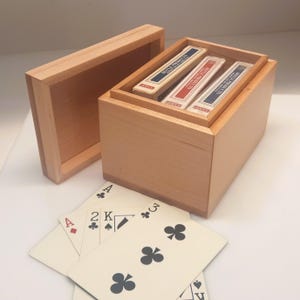 May include: A wooden box with a sliding lid, containing three decks of playing cards. The cards are in a wooden tray inside the box. Playing cards are fanned out in front of the box, showing the Ace of Diamonds, 2 of Clubs, King of Spades, and 3 of Clubs.