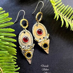 May include: A pair of artisan earrings with a gold and silver design. Each earring features a red stone set in an oval shape, with a gold ring above and a silver triangle below. The text "Dazzle Silver Stone" is in the lower right corner.