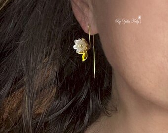 May include: A close-up of a gold earring with a white and yellow flower design. The earring is a drop style, with a long, thin gold bar. The flower has white petals and a yellow center. The text "By Yulia Kelly" is visible.