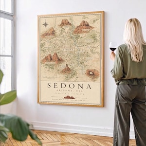 May include: A vertical wall art print featuring a detailed vintage-style map of Sedona, Arizona. The map displays red rock formations, roads, and landmarks with text labels. The print is framed in a light wood frame and mounted on a white wall.