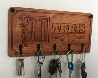 May include: Wooden key holder with the name "HARRIS" and the letter "H" engraved on it. The key holder has five hooks and is stained a dark reddish brown color.