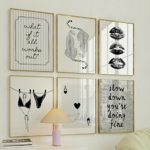 Può includere: Una collezione di stampe artistiche incorniciate con vari disegni. Le stampe includono testo, illustrazioni di leopardi, impronte di labbra e un disegno a tratteggio di bikini. Le cornici hanno bordi color oro. L'arte è esposta sopra un mobile bianco con una lampada.