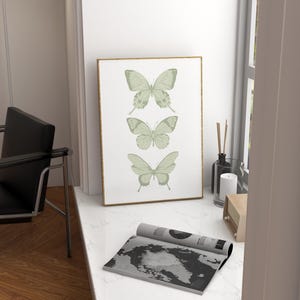 May include: A framed print featuring three green butterflies on a white background. The butterflies are arranged vertically, with the largest butterfly at the top and the smallest butterfly at the bottom. The frame is gold.