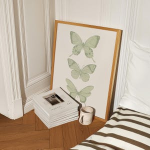 May include: A framed print with a pale green background featuring three stylised butterflies. The butterflies are arranged vertically, with the largest butterfly at the top and the smallest butterfly at the bottom. The frame is a light wood colour.