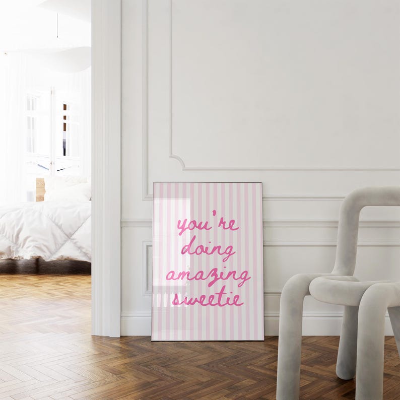 May include: A framed print with a pink and white striped background and the words "you're doing amazing sweetie" in pink script. The print is leaning against a white wall next to a gray chair.