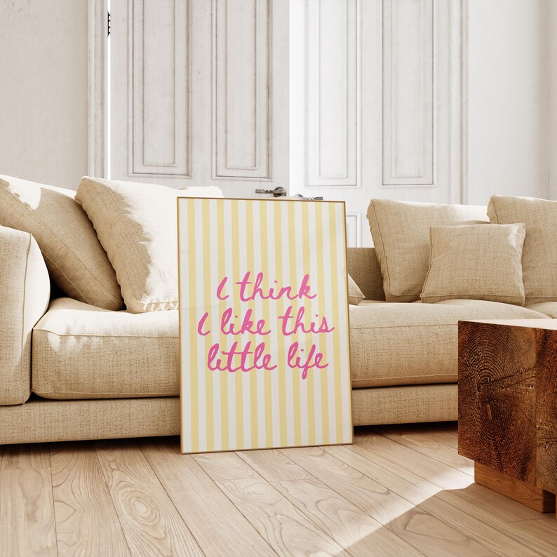 Puede incluir: Una impresi&oacute;n art&iacute;stica enmarcada con un fondo a rayas amarillas y blancas y el texto "I think I like this little life" en escritura rosa. La impresi&oacute;n se exhibe en un suelo de madera frente a un sof&aacute; beige.