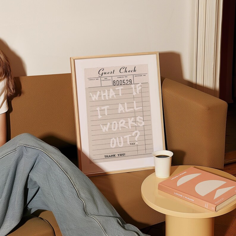 Puede incluir: Una impresi&oacute;n enmarcada de un "Guest Check" con el texto "WHAT IF IT ALL WORKS OUT?" en blanco. Una taza de caf&eacute; blanca peque&ntilde;a y un libro con una cubierta rosa est&aacute;n sobre una mesa peque&ntilde;a, redonda y amarilla. La impresi&oacute;n est&aacute; en un marco de madera clara.