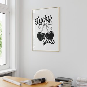 May include: Black and white print with the text "Lucky You" and a graphic of two cherries with a bow on top. The print is framed in a gold frame.