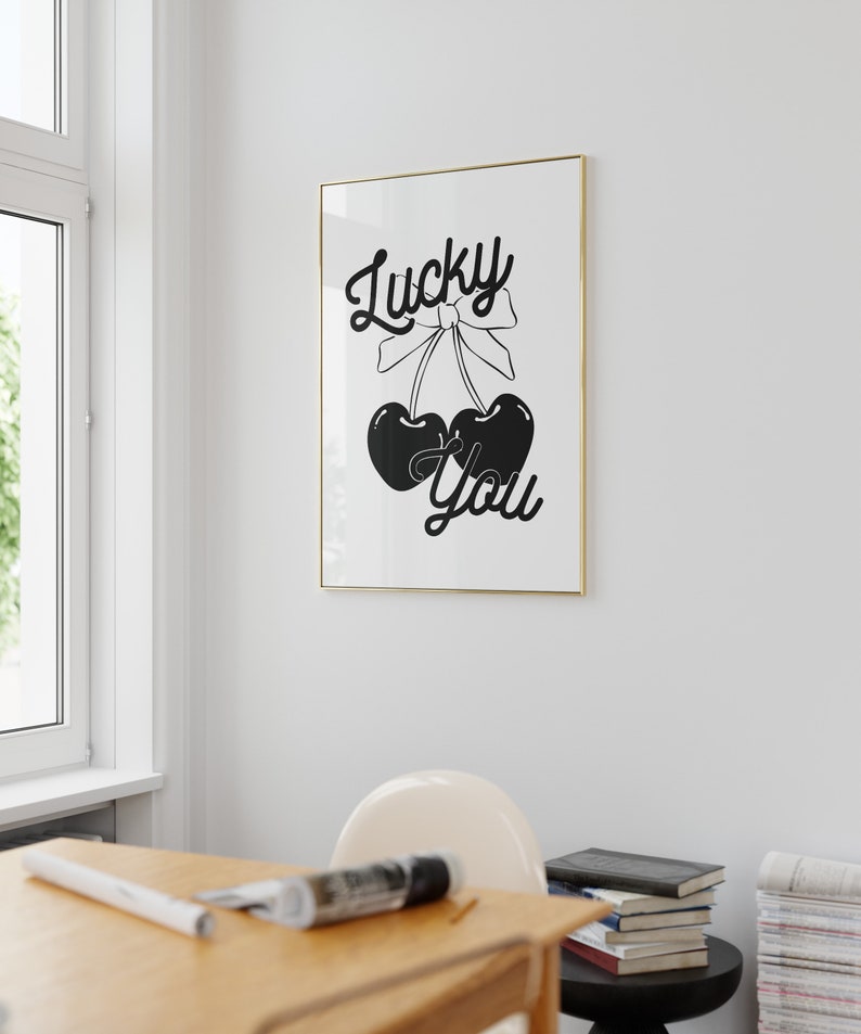 May include: Black and white print with the text "Lucky You" and a graphic of two cherries with a bow on top. The print is framed in a gold frame.