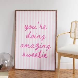 May include: A framed print with a pink and white striped background. The pink text reads "you're doing amazing sweetie". The frame is brown. A chair with a gold frame and a glass sphere are in the background.