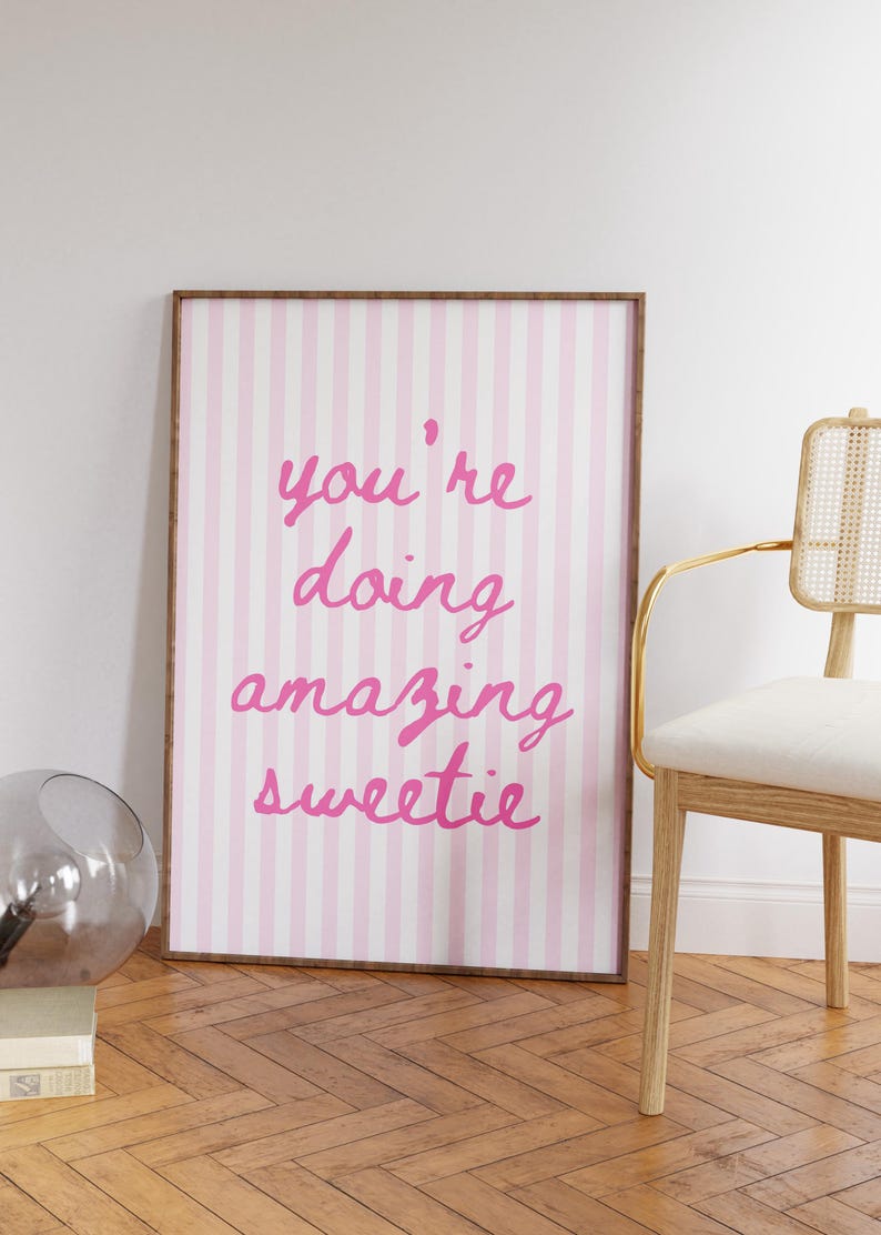 May include: A framed print with a pink and white striped background. The pink text reads "you're doing amazing sweetie". The frame is brown. A chair with a gold frame and a glass sphere are in the background.