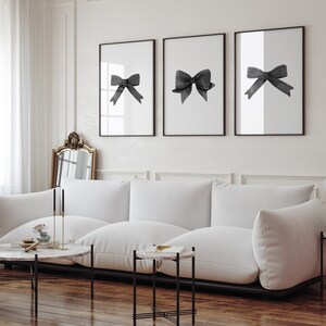 K&ouml;nnte beinhalten: Drei schwarz-wei&szlig;e gerahmte Drucke von schwarzen Aquarell-Schleifen auf wei&szlig;em Hintergrund. Die Drucke h&auml;ngen an einer wei&szlig;en Wand &uuml;ber einem wei&szlig;en Sofa.