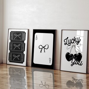 May include: Three black and white framed prints with different designs. The left print features three ticket stubs with a bow on each. The middle print features a playing card with a black bow on a white background. The right print features two cherries with a bow and the text "Lucky You".