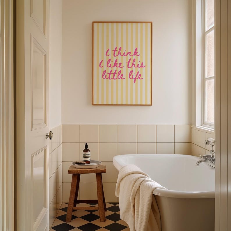 Puede incluir: Interior de ba&ntilde;o con un estampado enmarcado con el texto "I think I like this little life" en escritura rosa sobre un fondo a rayas amarillas. Un taburete de madera sostiene una bandeja con una botella y un cepillo. Tambi&eacute;n hay una ba&ntilde;era blanca.