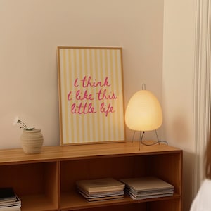Puede incluir: Un estampado enmarcado con rayas amarillas y blancas y el texto "I think I like this little life" en rosa. Una l&aacute;mpara de papel blanco con patas negras y un peque&ntilde;o jarr&oacute;n con flores blancas se encuentran en una estanter&iacute;a de madera.