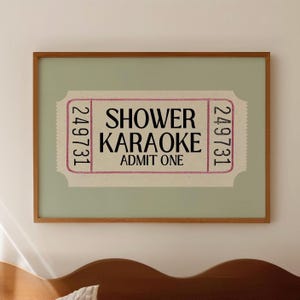May include: A framed print featuring a vintage-style ticket design. The ticket reads "Shower Karaoke, Admit One" in black text, with the number "249731" on each side. The frame is a warm brown color, and the background is a soft green.