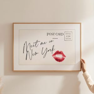 Pode incluir: Uma impressão emoldurada de cartão postal com o texto "Meet me in New York" em escrita elegante. A impressão apresenta uma marca de beijo de batom vermelho e um design de cartão postal vintage. A moldura é de madeira clara.
