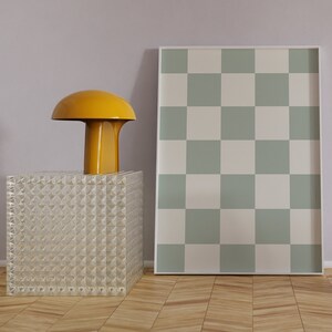 Puede incluir: Una l&aacute;mpara amarilla con forma de hongo se encuentra sobre una mesa de cubo de vidrio transparente frente a una impresi&oacute;n a cuadros verde y blanca enmarcada.