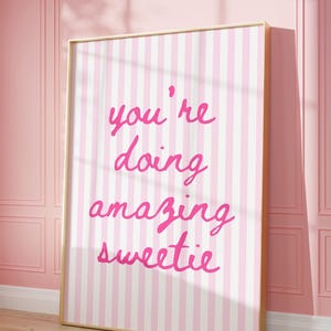 May include: A framed print with a pink and white striped background. The print features the handwritten text "you're doing amazing sweetie" in bright pink. The frame is a light wood color, and the print is leaning against a pink wall.