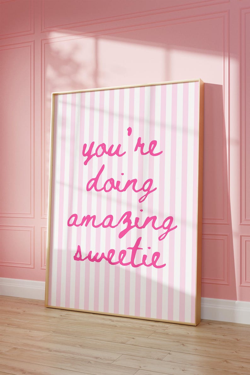 May include: A framed print with a pink and white striped background. The print features the handwritten text "you're doing amazing sweetie" in bright pink. The frame is a light wood color, and the print is leaning against a pink wall.