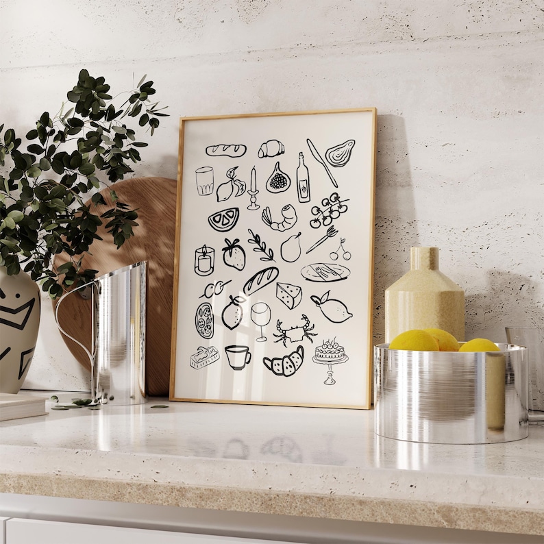 May include: A black and white illustration of various food and drink items, including a baguette, grapes, a glass of wine, a lemon, a crab, and a fig. The illustration is framed in a light wood frame and is hanging on a wall.