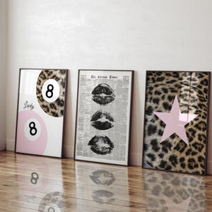 May include: Three framed art prints leaning against a wall. One features a leopard print eight ball design with the word "Lucky" and the number 8. The second print has three lip prints. The third print has a pink star.
