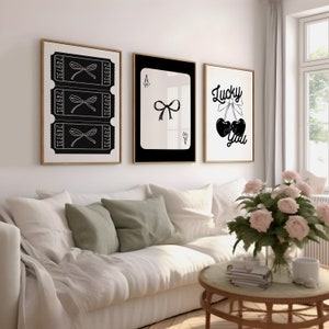 May include: Three black and white framed prints on a white wall above a white couch with green pillows. The prints feature a black bow, a playing card with a black bow, and a black cherry with a bow and the text "Lucky You".