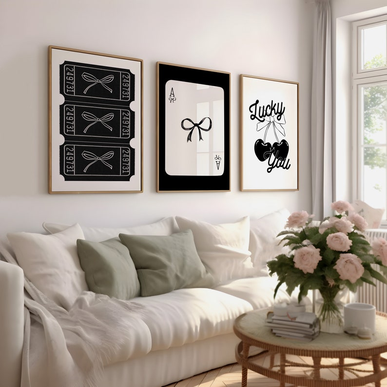 May include: Three black and white framed prints on a white wall above a white couch with green pillows. The prints feature a black bow, a playing card with a black bow, and a black cherry with a bow and the text "Lucky You".