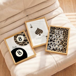 May include: Three gold-framed wall art prints. One features two billiard 8-balls, one with a leopard print pattern and the word Lucky. Another shows an ace of spades with a leopard print design and the text Feeling Lucky. The third has leopard print with the text YOU LOOK SO GOOD.