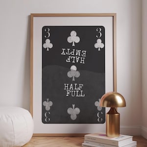 Op de afbeelding: Ingelijste kunstdruk van een speelkaart, de drie van klaveren, met de woorden "Half Empty" boven "Half Full" in witte tekst op een zwarte achtergrond. De print zit in een houten lijst.