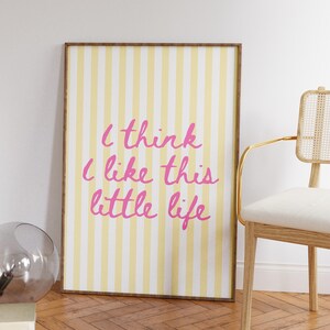 Puede incluir: Un estampado enmarcado con un fondo a rayas amarillas y blancas y el texto rosa "I think I like this little life". El estampado est&aacute; en un marco marr&oacute;n y se apoya en una pared blanca. Una silla con marco dorado y un jarr&oacute;n de cristal est&aacute;n cerca.