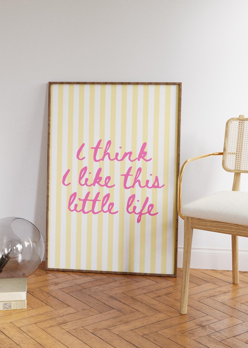 Puede incluir: Un estampado enmarcado con un fondo a rayas amarillas y blancas y el texto rosa "I think I like this little life". El estampado est&aacute; en un marco marr&oacute;n y se apoya en una pared blanca. Una silla con marco dorado y un jarr&oacute;n de cristal est&aacute;n cerca.