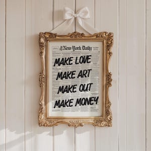 以下が含まれることがあります： The New York Dailyというタイトルのヴィンテージ新聞ページに、黒い文字でMAKE LOVE, MAKE ART, MAKE OUT, MAKE MONEYと印刷された額入りプリント。フレームは装飾が施されたゴールドで、上部中央に白いリボンの結び目が付いています。
