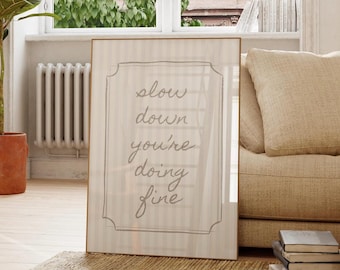 May include: A framed print with the words "slow down you're doing fine" in a handwritten style. The print has a light beige striped background and a thin, light brown frame. It is leaning against a wall near a window and a beige sofa.