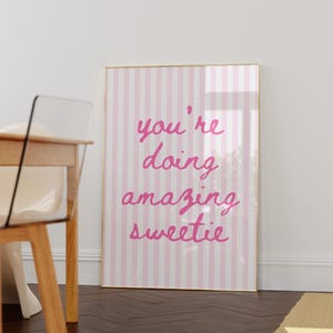 May include: A framed print with a pink and white striped background. The print features the handwritten text "you're doing amazing sweetie" in a bright pink color. The frame is gold.