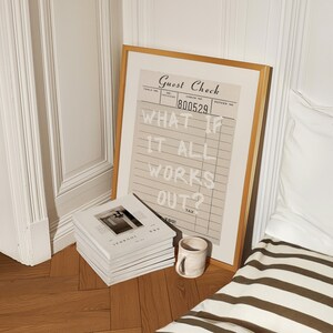 Puede incluir: Una impresi&oacute;n enmarcada con el texto "WHAT IF IT ALL WORKS OUT?" se encuentra en un suelo de madera junto a una pila de revistas y una taza peque&ntilde;a. La impresi&oacute;n se asemeja a un recibo de restaurante, con un fondo beige y un marco de madera. Las revistas son blancas.