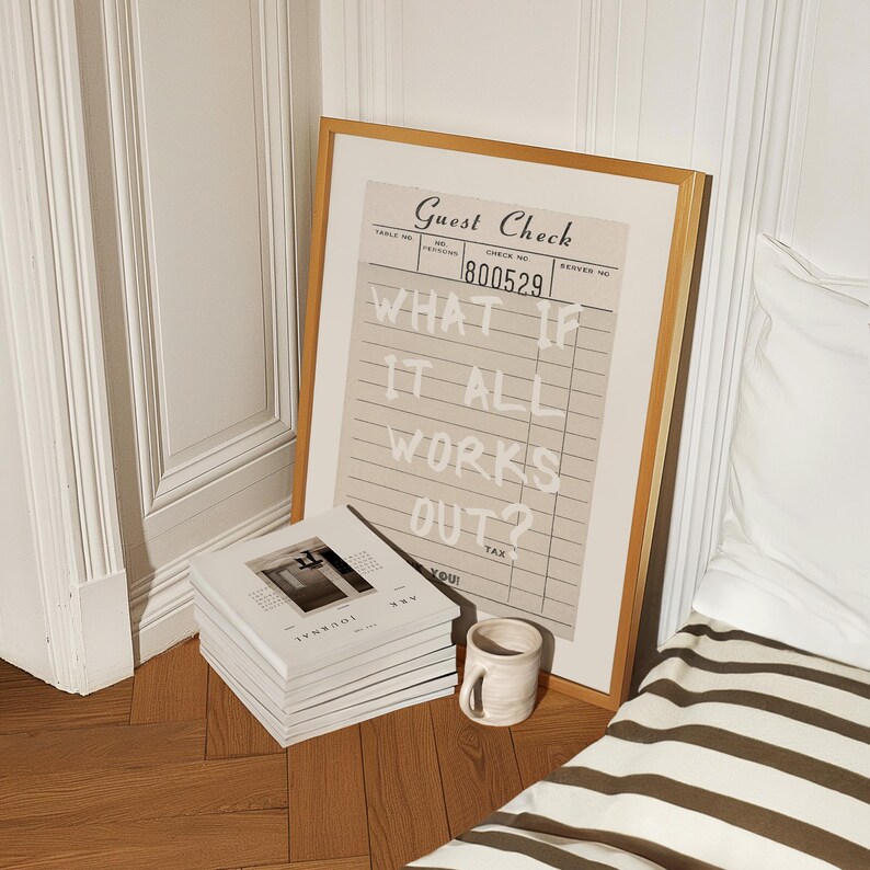 Puede incluir: Una impresi&oacute;n enmarcada con el texto "WHAT IF IT ALL WORKS OUT?" se encuentra en un suelo de madera junto a una pila de revistas y una taza peque&ntilde;a. La impresi&oacute;n se asemeja a un recibo de restaurante, con un fondo beige y un marco de madera. Las revistas son blancas.