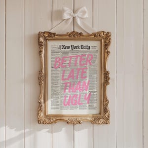 May include: A framed print of The New York Daily newspaper featuring the pink text BETTER LATE THAN UGLY overlaid on the center. The print is housed in an ornate gold frame with a white ribbon bow attached to the top for hanging display.