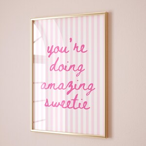 May include: A framed print with a gold-colored border. The artwork features a pink and white striped background with the handwritten phrase "you're doing amazing sweetie" in bright pink lettering.